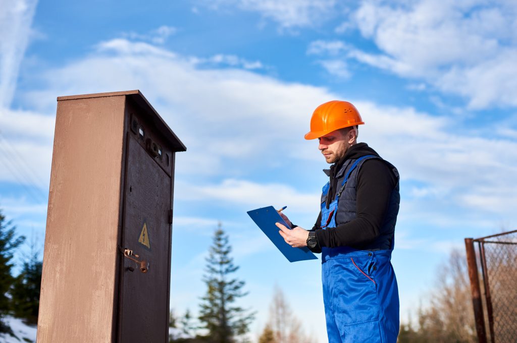 Oil inspector in protective overalls and orange helmet making notes next to a transformer outdoors against blue sky with white clouds. Concept of petroleum industry.